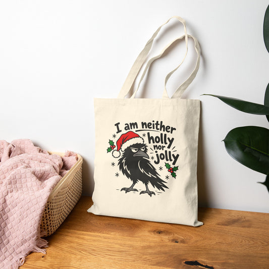 Natural canvas tote bag with a grumpy raven in a Santa hat, holly, and 'I am neither holly nor jolly' text, standing on a wood table beside a basket and pink knit blanket.