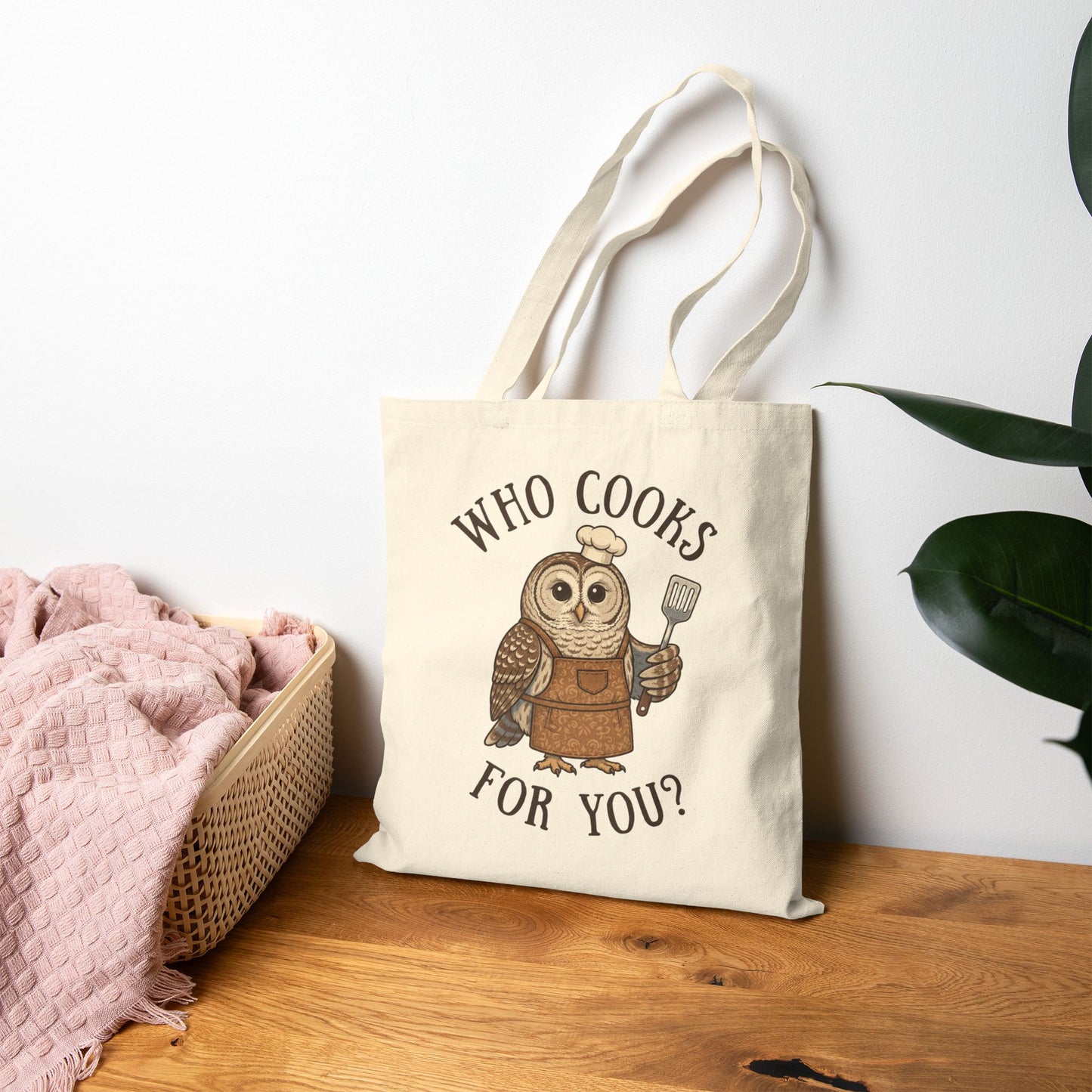 A cream canvas tote bag with a printed cartoon owl chef holding a spatula, with the text 'WHO COOKS FOR YOU?'. The bag is styled on a wooden table next to a pink blanket in a basket and a green plant against a white wall.