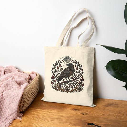 Tote bag with a raven design on a wooden surface next to a pink blanket and plant.