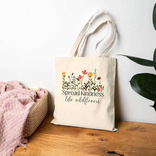 Tote bag with floral design and 'Spread Kindness like wildflowers' text on a wooden surface.