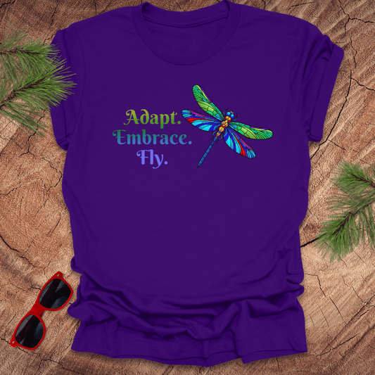 Purple t-shirt with a colorful dragonfly graphic and "adapt. embrace. fly. text on a wooden surface with sunglasses and greenery.