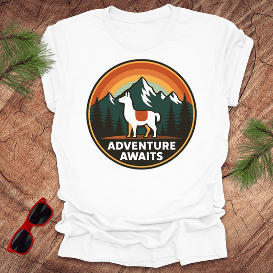 White t-shirt with 'Adventure Awaits' graphic featuring a llama and mountains on a wooden surface with sunglasses and pine branches.