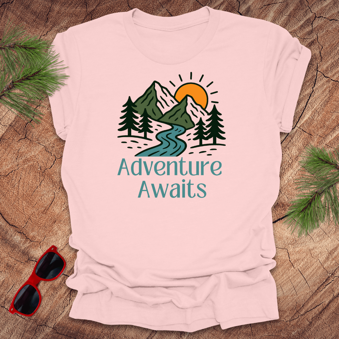 Pink t-shirt with 'Adventure Awaits' graphic on a wooden surface with sunglasses and pine branches.