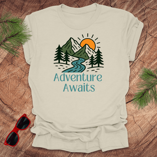 Beige t-shirt with 'Adventure Awaits' graphic on a wooden surface with sunglasses and pine branches.
