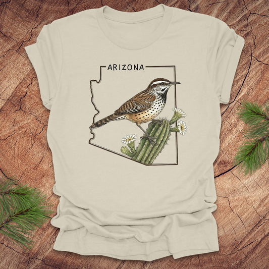 Beige t-shirt with Arizona state outline with cactus wren and cactus flower design on a wooden surface