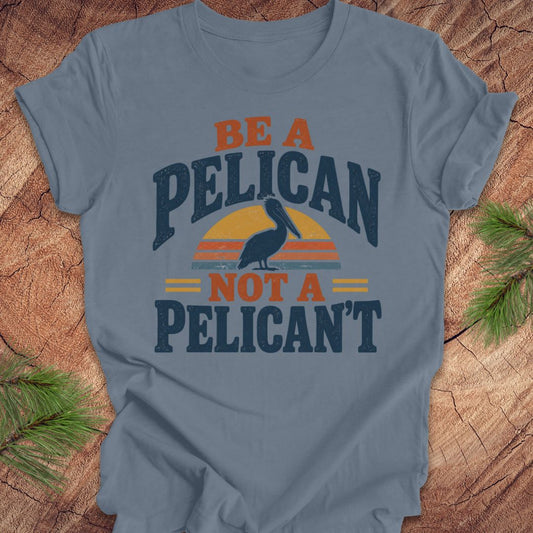 Steel blue t-shirt with a Brown Pelican silhouette on a retro striped sunset and the words BE A PELICAN NOT A PELICAN'T, laid flat on a rustic wood background with pine branches.