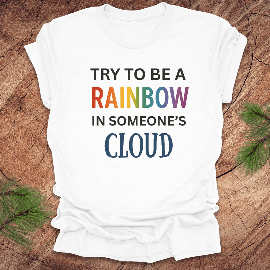 White t-shirt with colorful text 'Try to be a rainbow in someone's cloud' on a wooden surface