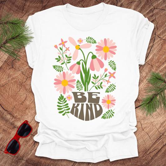 White t-shirt with floral design and 'Be Kind' text on a wooden surface with sunglasses and greenery.