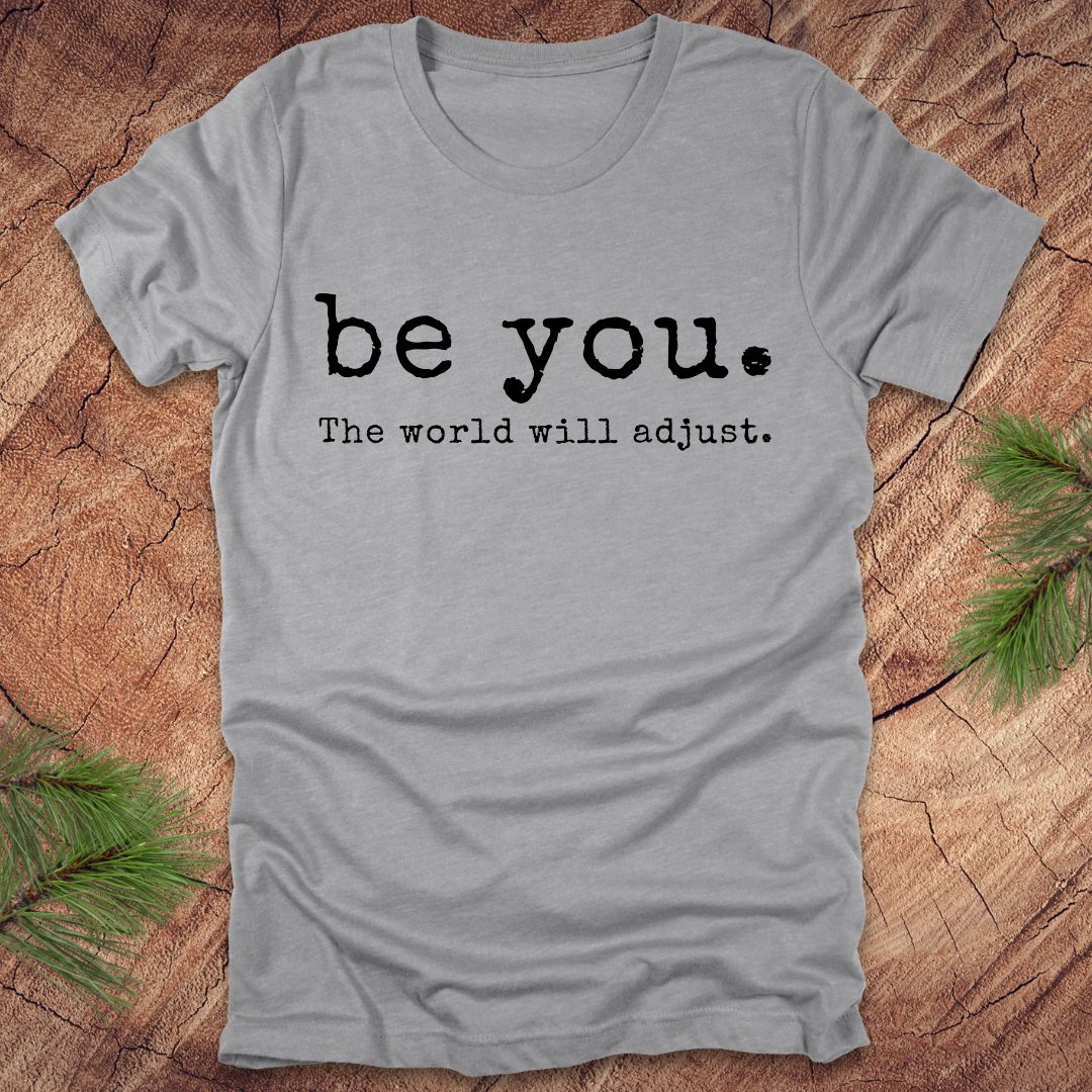 Gray t-shirt with 'be you. The world will adjust.' text on a wooden surface
