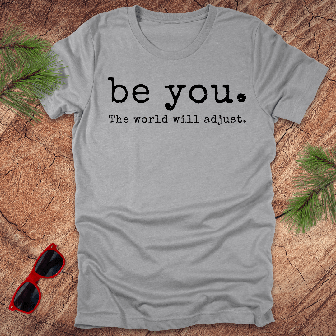 grey heather tshirt with the text Be you. the world will adjust on a wood background with sunglasses and pine
