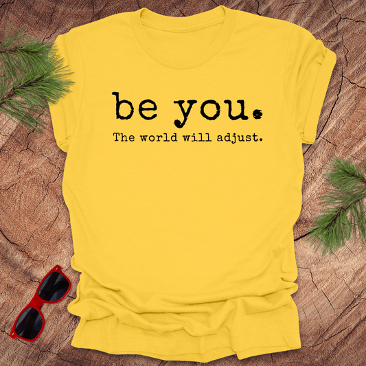 yellow tshirt with the text Be you. the world will adjust on a wood background with sunglasses and pine