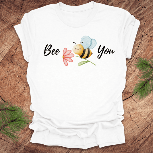 White tshirt with a cute bee and the text, "bee you" on a wooden background.