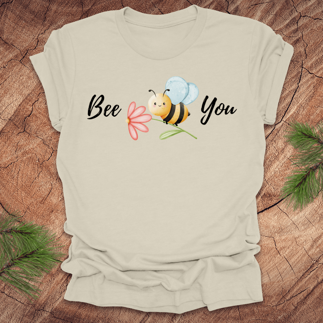 Beige tshirt with a cute bee and the text, "bee you" on a wooden background.