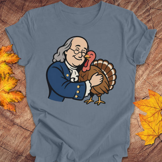 A slate blue short-sleeve t-shirt laid flat on a rustic wood background with autumn leaves. The front of the shirt features a cartoon graphic of Benjamin Franklin hugging a brown turkey.