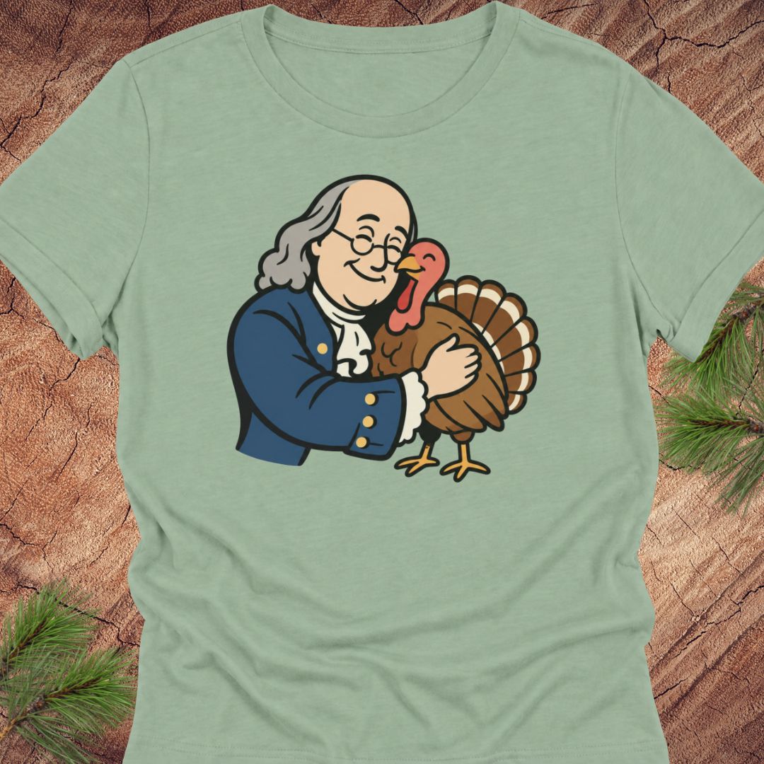 A sage green short-sleeve t-shirt featuring a cartoon graphic of George Washington hugging a turkey, displayed flat on a rustic wood background with pine needles.