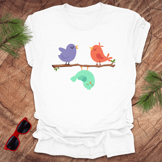 White t-shirt with colorful birds design on a wooden surface with sunglasses and greenery.
