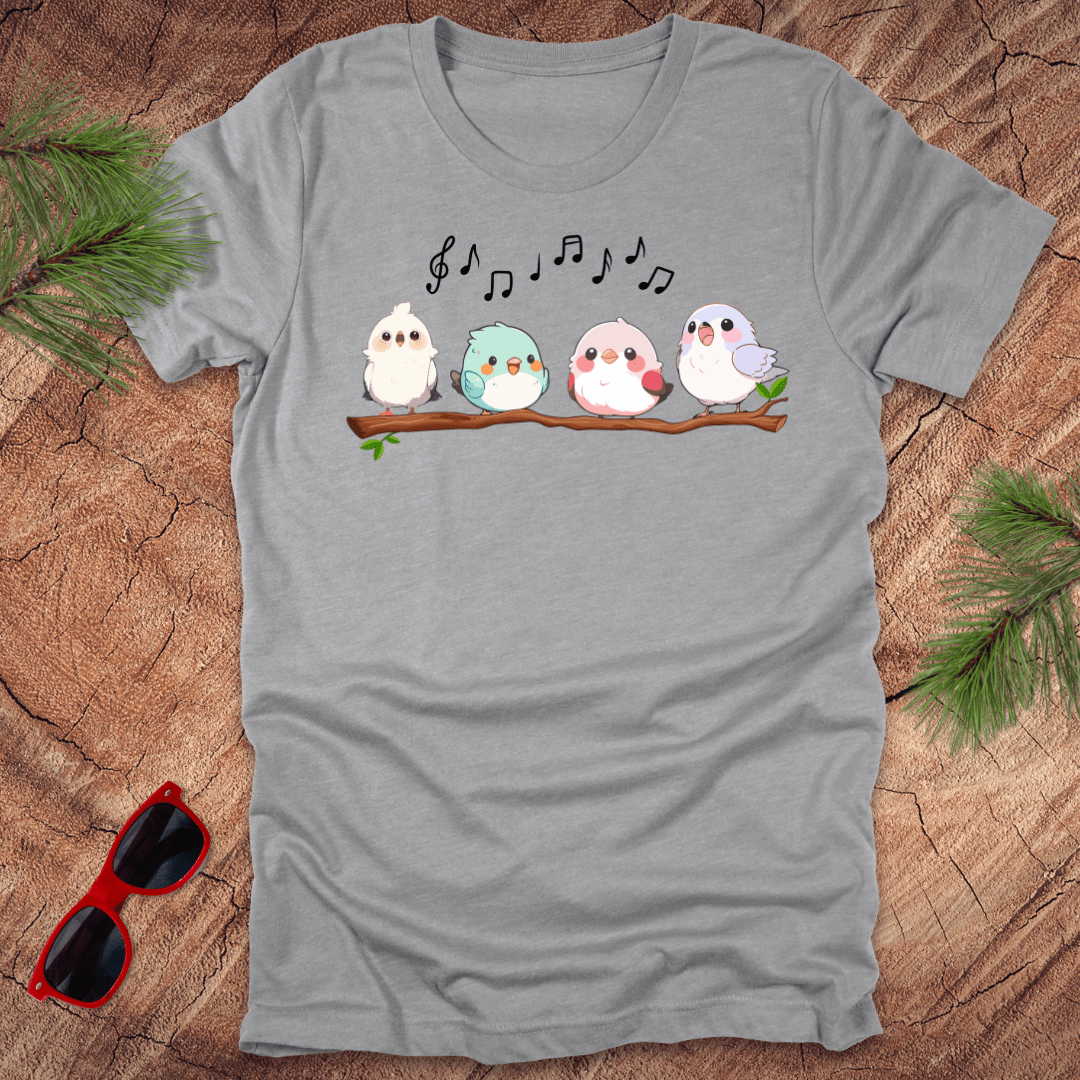 Gray t-shirt with cartoon birds and musical notes on a wooden surface with sunglasses and greenery.