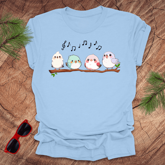 Light blue t-shirt with cartoon birds and musical notes on a wooden surface with sunglasses and greenery.