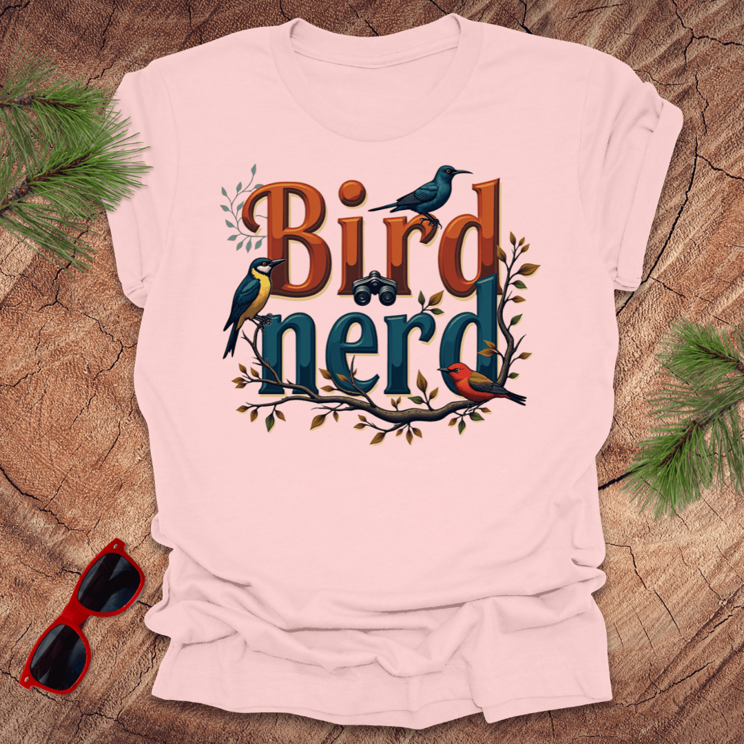 A pink t-shirt with a graphic print of birds and the text 'Bird nerd', surrounded by foliage and a pair of sunglasses lying next to it on a wooden surface.