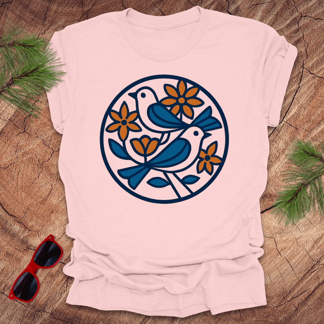 Pink t-shirt with a blue and orange bird and flower design on a wooden surface with sunglasses and greenery.