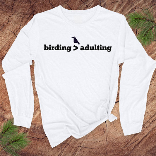 White long-sleeve shirt with 'birding > adulting' text on a wooden surface with greenery.