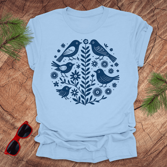 Light blue t-shirt with bird and floral design on a wooden surface with sunglasses and greenery.