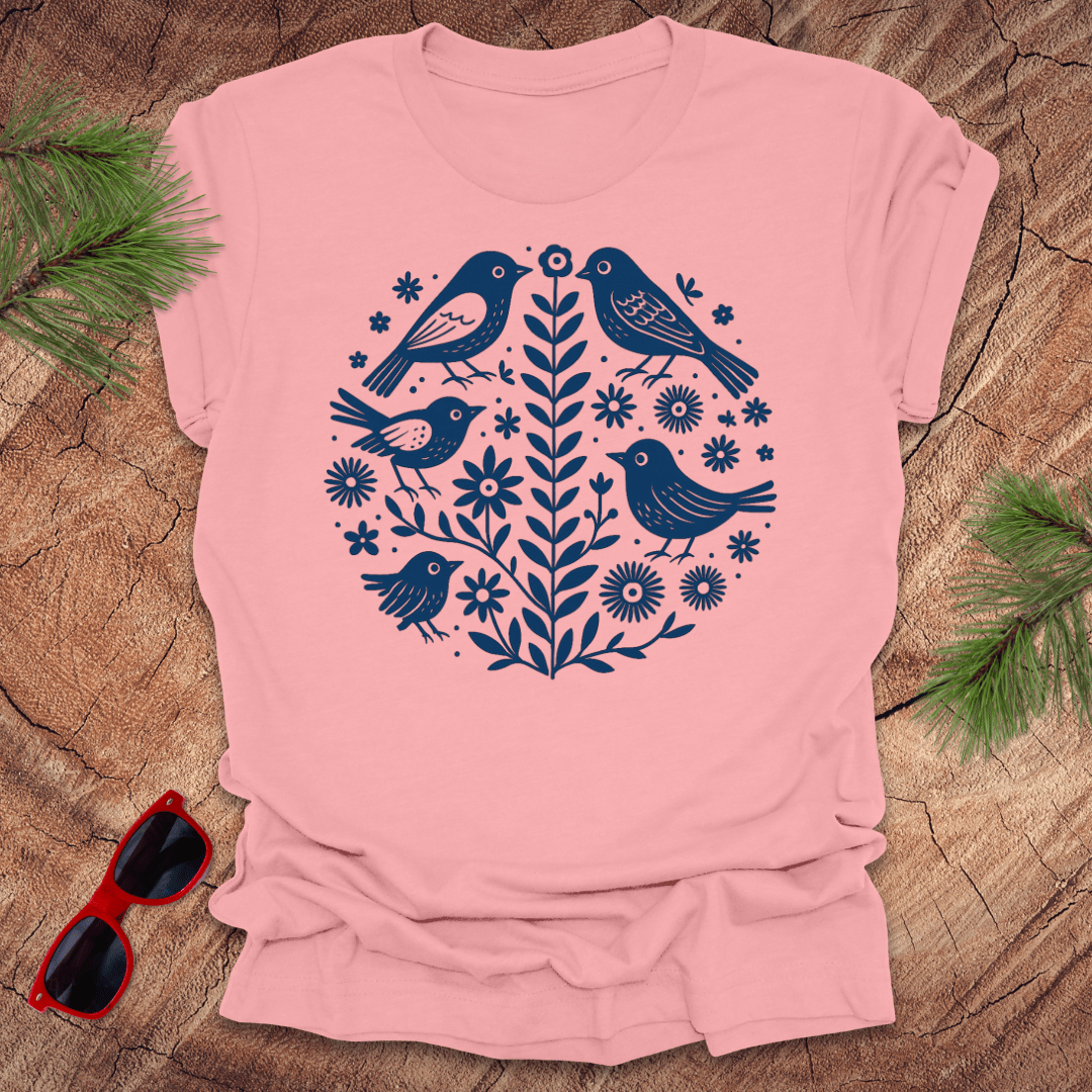 Pink t-shirt with blue bird and floral design on a wooden surface with sunglasses and greenery.