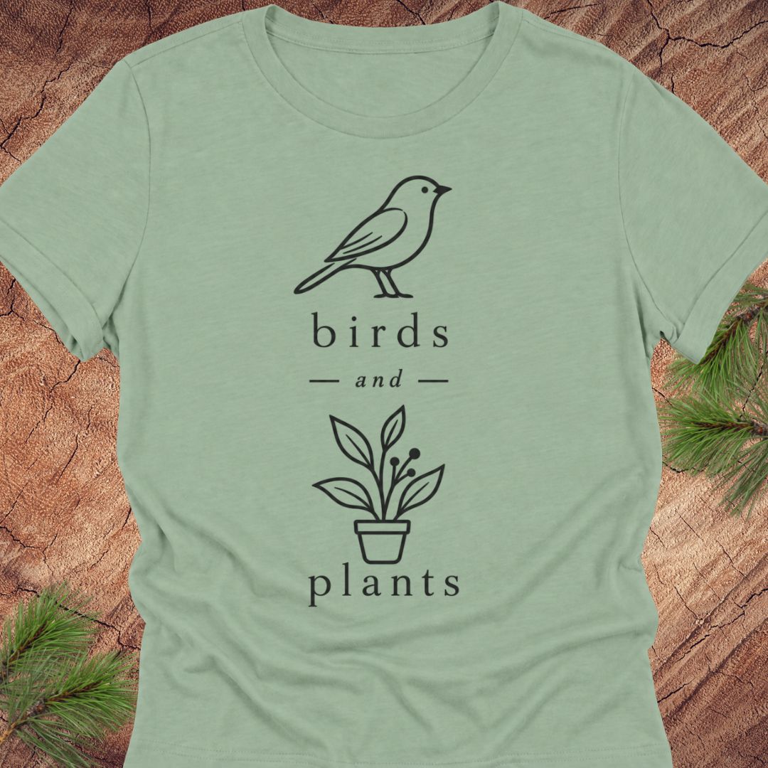 A sage green short-sleeve t-shirt with a black line art graphic of a bird and a potted plant, with the words 'birds and plants' printed below. The shirt is laid flat on a rustic wood background with pine branches in the corners.