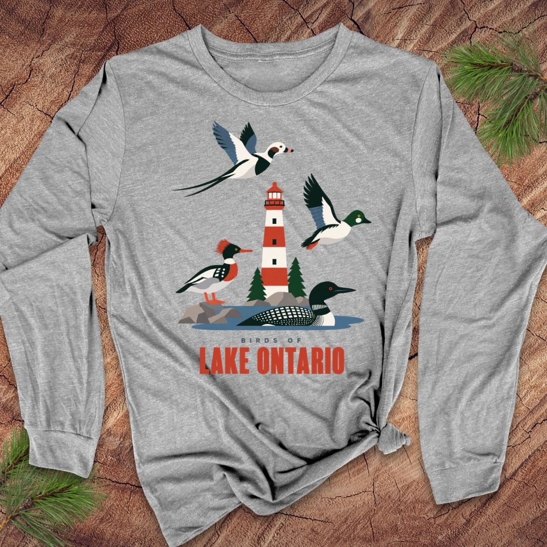 A heather gray long-sleeve t-shirt with a colorful graphic of birds and a lighthouse around Lake Ontario, displayed flat on a rustic wood background with pine needles.