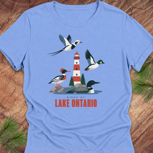 Carolina blue women's relaxed t-shirt with a red-and-white lighthouse, loon, merganser, goldeneye, and long-tailed duck above 'Birds of Lake Ontario' text, laid flat on a rustic wood background with pine branches.