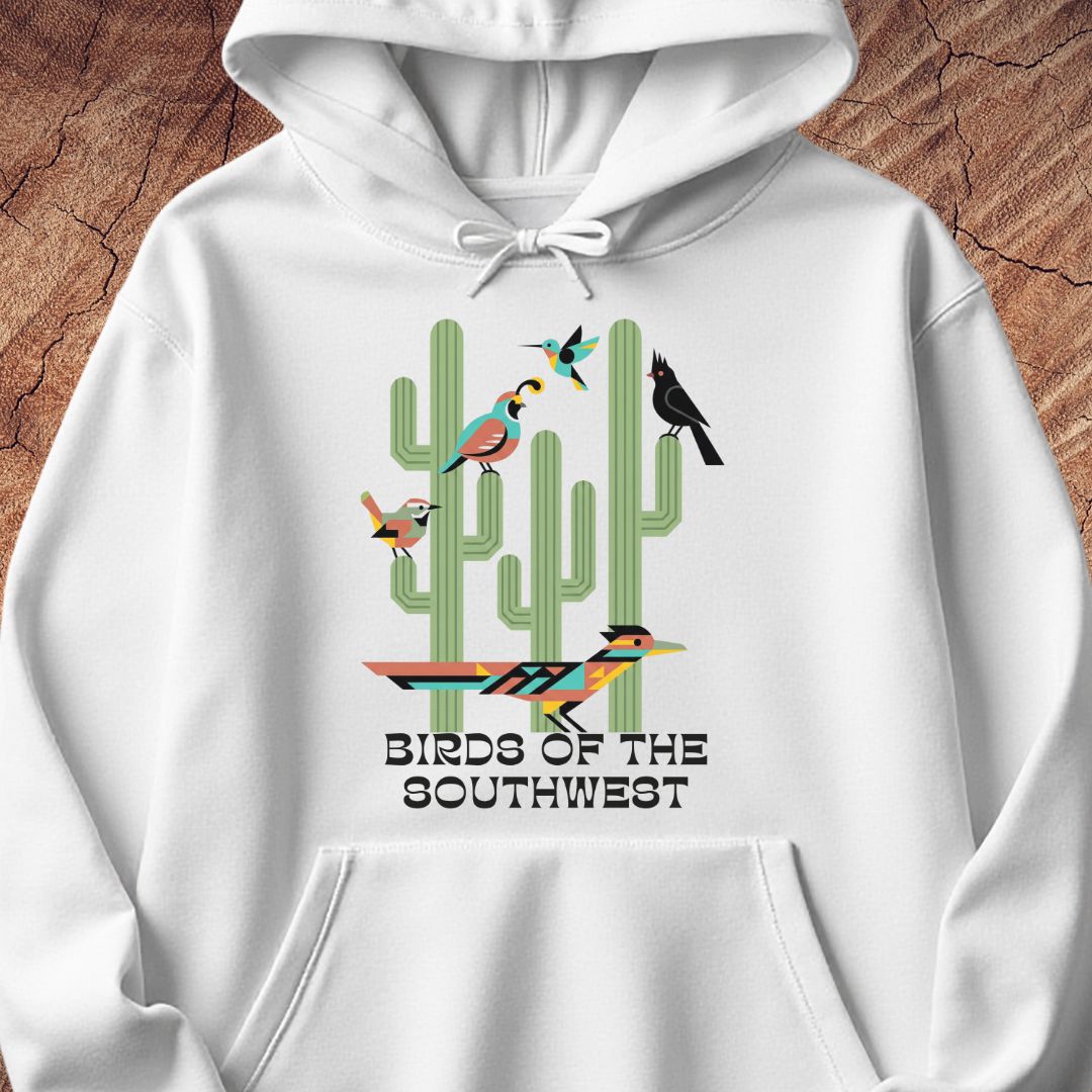 A white pullover hoodie with a colorful graphic print of cacti and birds, featuring the text 'BIRDS OF THE SOUTHWEST' underneath, displayed on a textured brown wood background.
