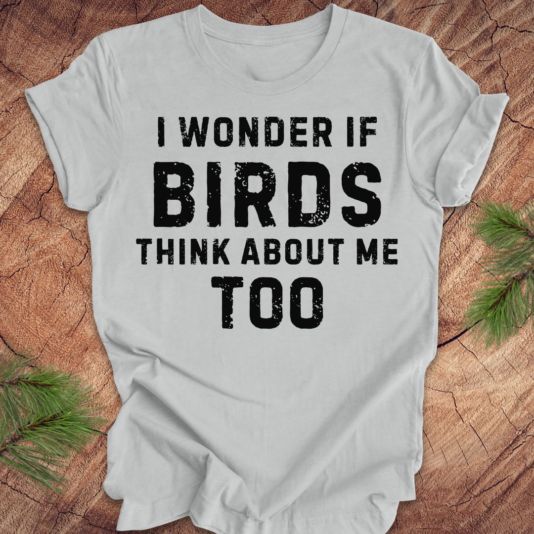 Gray t-shirt with black text 'I wonder if birds think about me too' on a wooden surface with greenery.