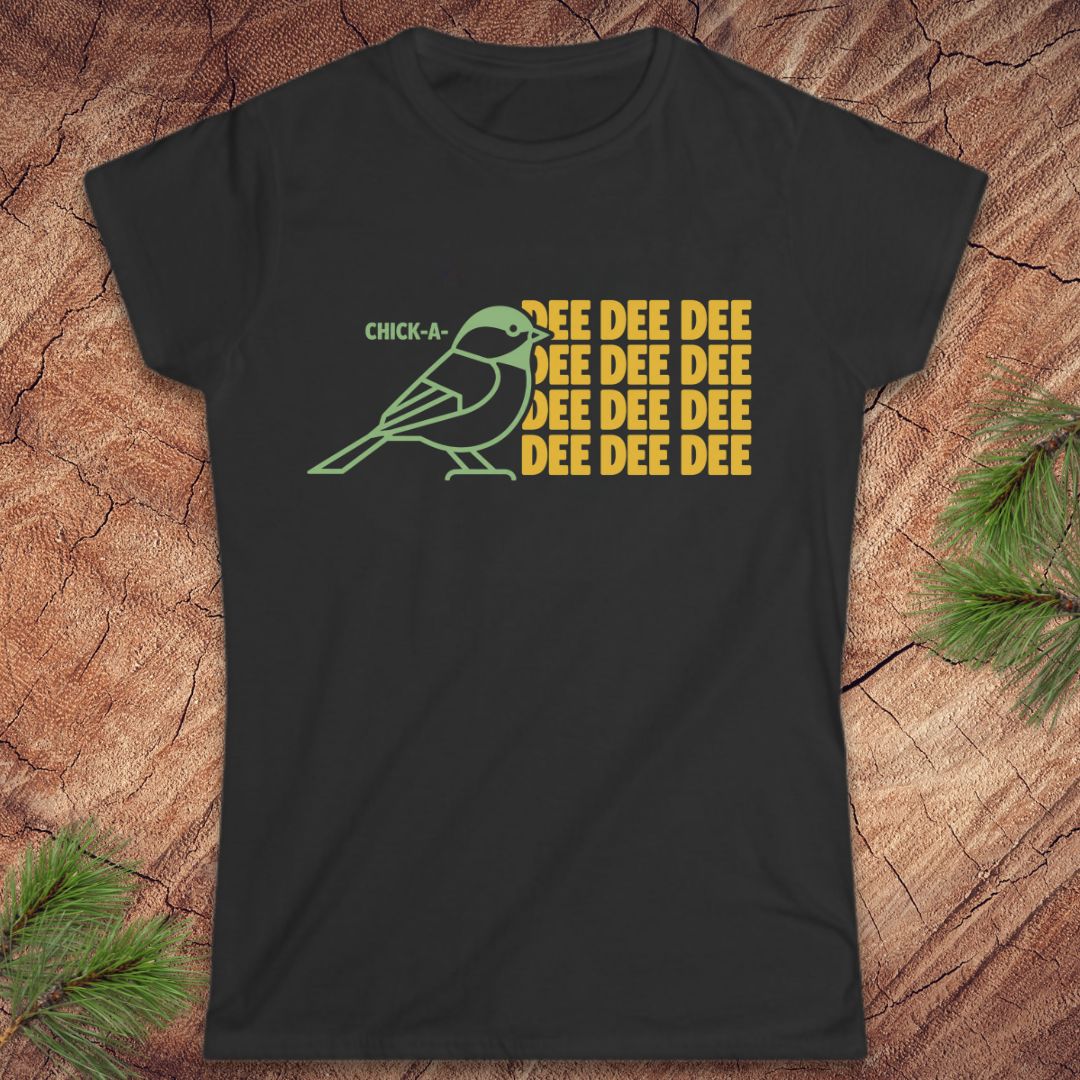 A black short-sleeve t-shirt with a graphic of a green chickadee and yellow text reading 'CHICK-A- DEE DEE DEE DEE DEE DEE', laid flat on a wood surface with pine branches in the corners.