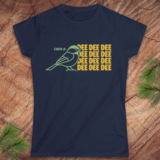 A navy blue short-sleeve t-shirt with a graphic of a green chickadee and yellow text reading 'CHICK-A- DEE DEE DEE DEE DEE DEE', laid flat on a wood surface with pine branches in the corners.