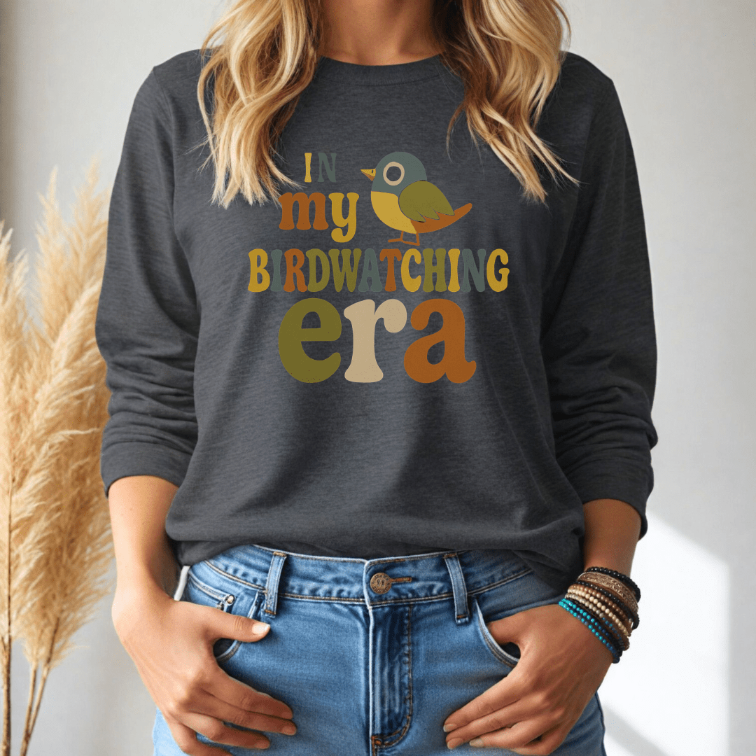 Person wearing a dark gray long-sleeve tshirt with colorful text 'In my birdwatching era' and a bird graphic, standing against a neutral background.