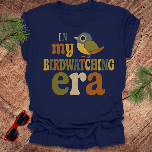 Navy blue t-shirt with colorful text and bird graphic on a wooden surface with sunglasses and pine branches.