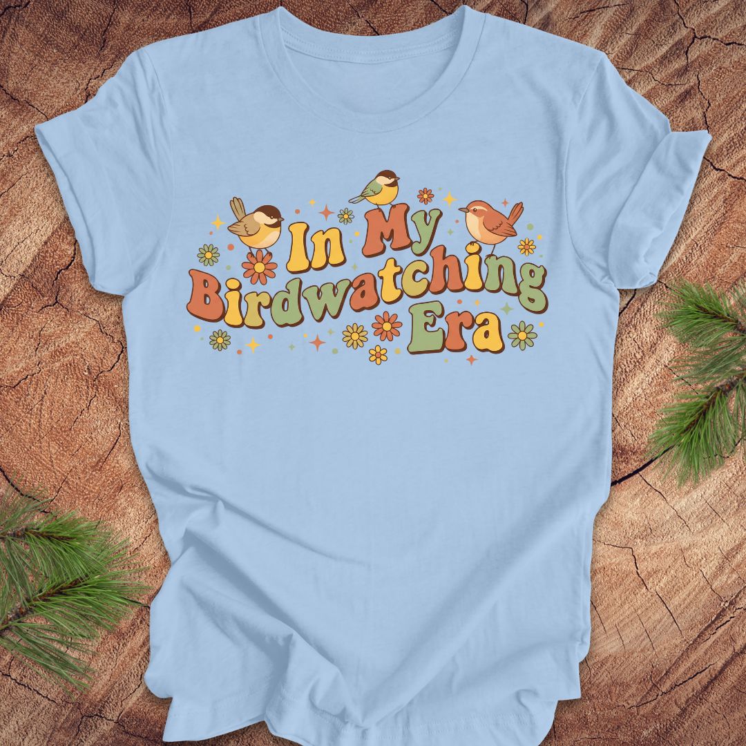 Light blue t-shirt with 'In My Birdwatching Era' text and bird illustrations on a wooden surface.