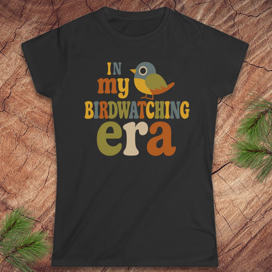 A black short-sleeve graphic t-shirt laid flat on a textured wood background with pine branches in the corners. The graphic reads 'IN my BIRDWATCHING era' in a retro multicolor font, accompanied by a small bird illustration.