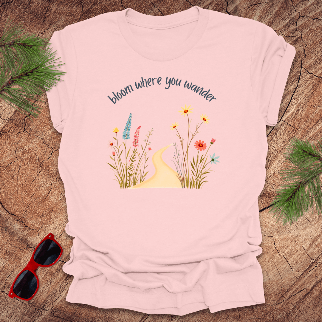 pink t-shirt with a graphic that includes a floral design and the text 'bloom where you wander', accompanied by  flowers on a wooden background with pine needles and red sunglasses to the side.