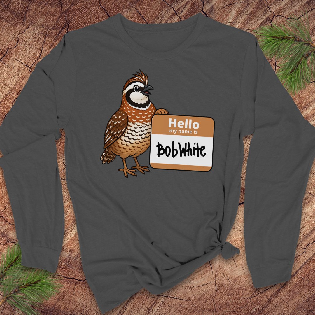 A dark gray long-sleeve t-shirt with a graphic of a brown and white quail next to a 'Hello My Name Is Bob White' nametag, displayed flat on a rustic wood background with pine needles.