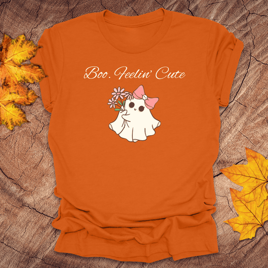 Orange t-shirt with a ghost wearing a pink bow and flowers, text 'Boo. Feelin' Cute', on a wooden surface with autumn leaves.