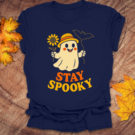 Navy blue t-shirt with a ghost holding a flower and 'Stay Spooky' text on a wooden surface with leaves.