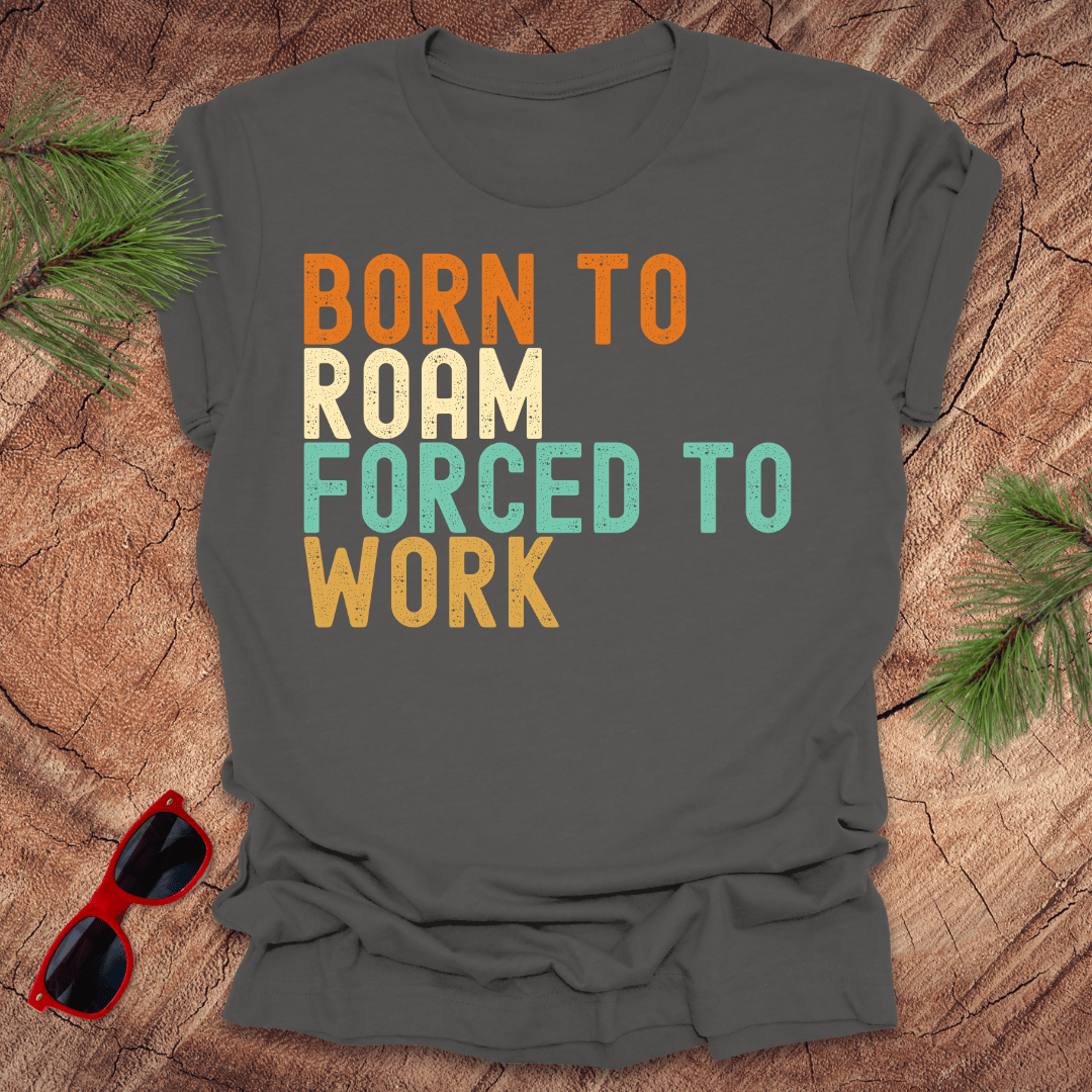silver tshirt with text "born to roam forced to work' on a wood background with pine needles and sunglasses