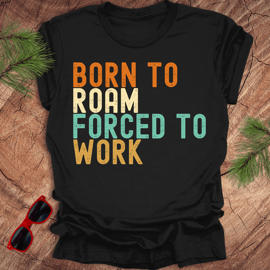 black tshirt with text "born to roam forced to work' on a wood background with pine needles and sunglasses