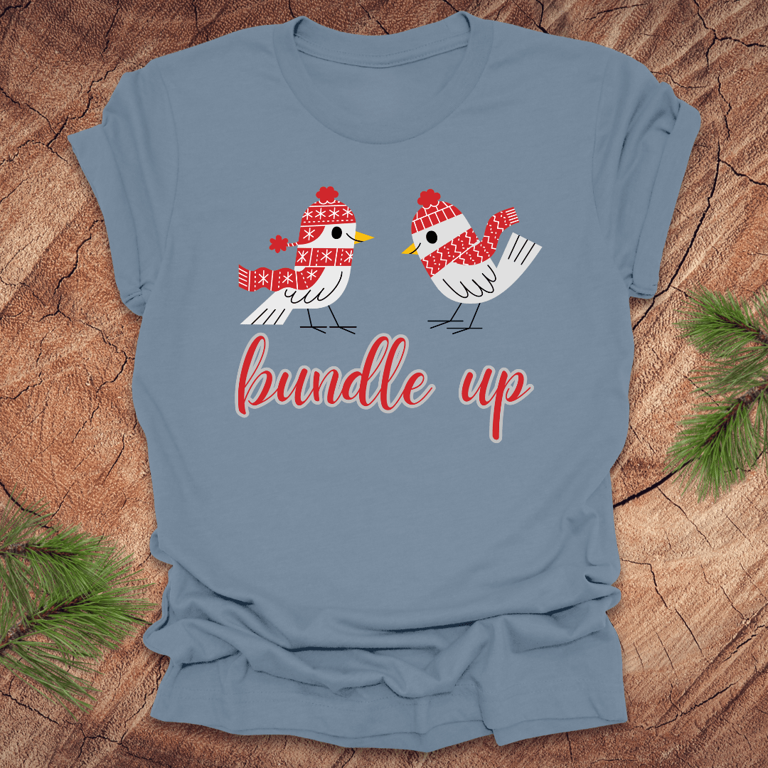 Light blue t-shirt with two birds with hats and scarves and text, 'Bundle up' on a wood background