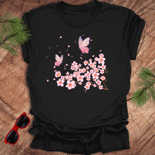 Black t-shirt with pink butterfly and cherry blossom design on a wooden surface with sunglasses and greenery.