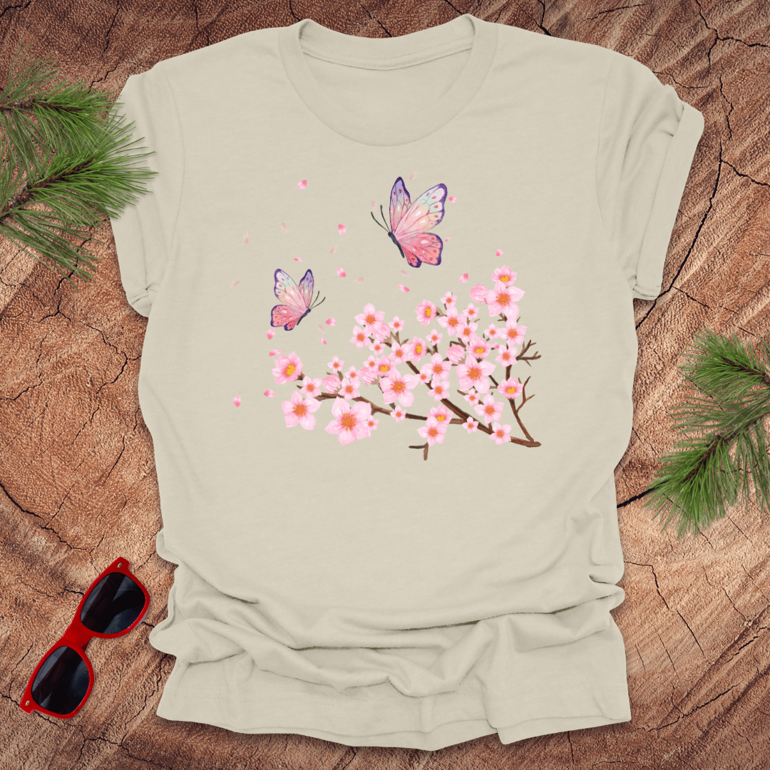Beige t-shirt with cherry blossom and butterfly design on a wooden surface with sunglasses and greenery.