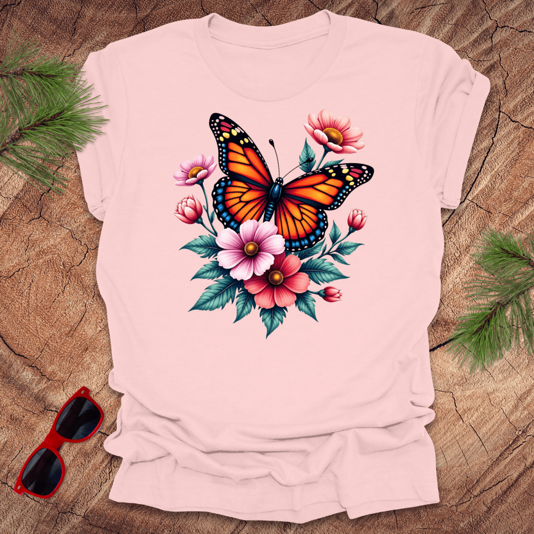 A pink t-shirt with a vibrant print of a butterfly surrounded by pink and red flowers, displayed on a wooden surface with green leaves and red sunglasses to the side.
