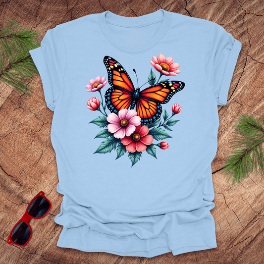 A baby blue t-shirt with a vibrant print of a butterfly surrounded by pink and red flowers, displayed on a wooden surface with green leaves and red sunglasses to the side.
