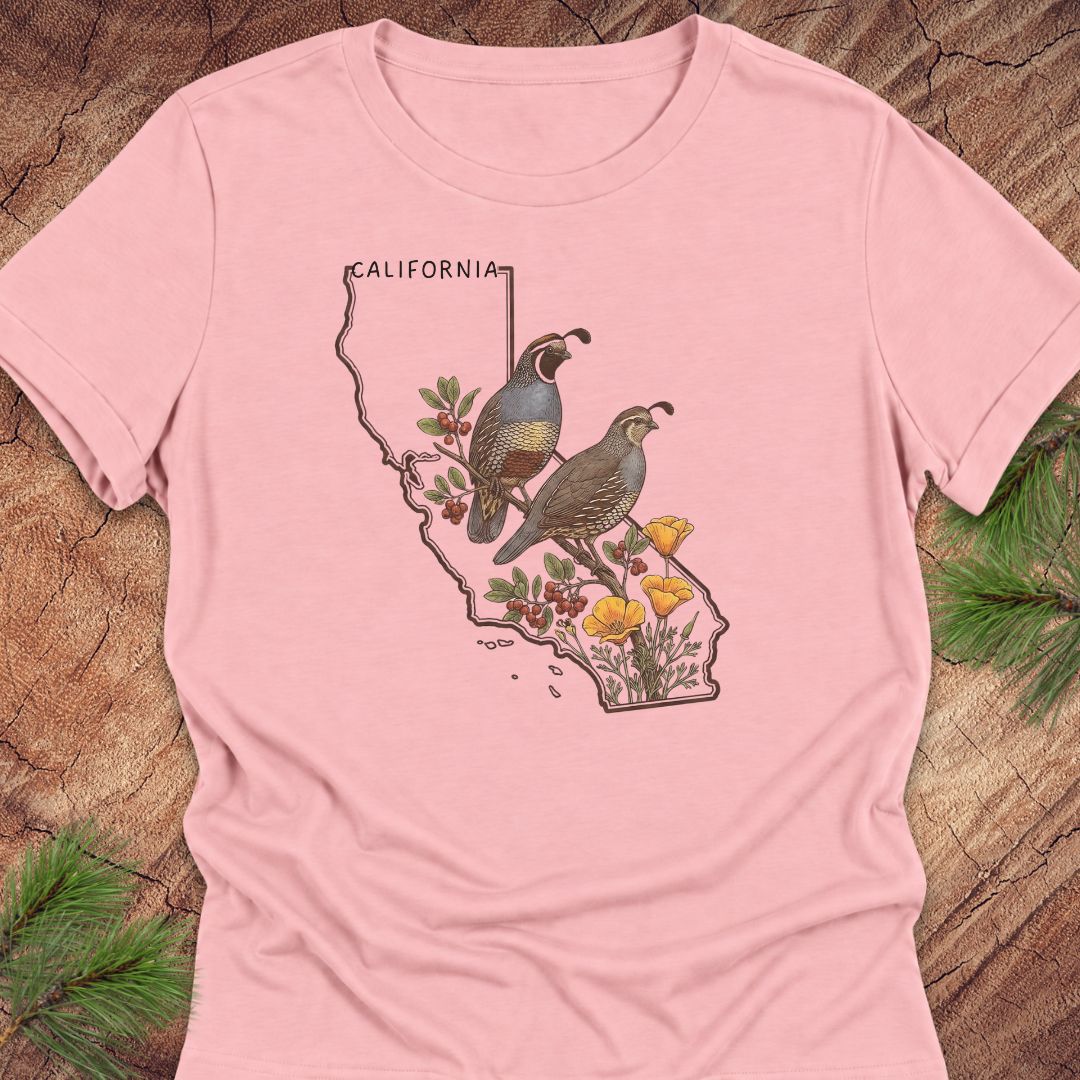 A light pink short-sleeve t-shirt with a graphic print of a California map outline featuring two quail birds, cactus plants, and yellow flowers. The shirt is laid flat on a rustic wood background with pine needles visible in the corners.